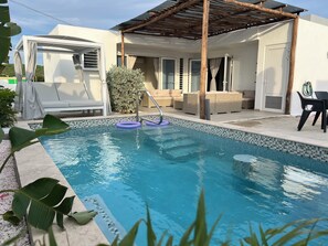 A swimming pool with a tiled edge, surrounded by lounge chairs and a covered patio area. - Casa Wechi: 3-Bedroom Central Pool Home For Families in a Secured Neighbourhood (Willemstad)