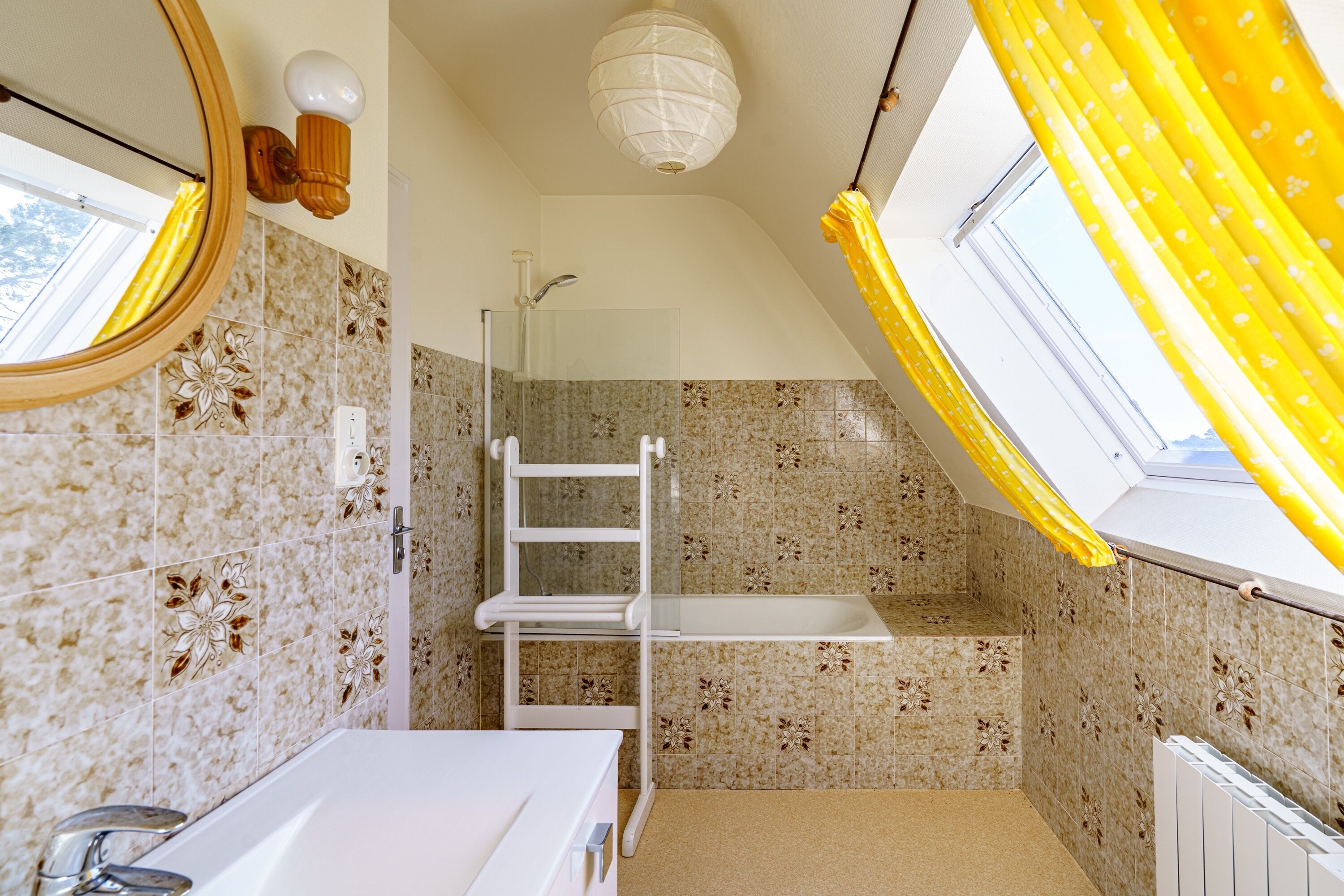 A bathroom with a shower, a white bathtub, a mirror, and a skylight with yellow curtains.