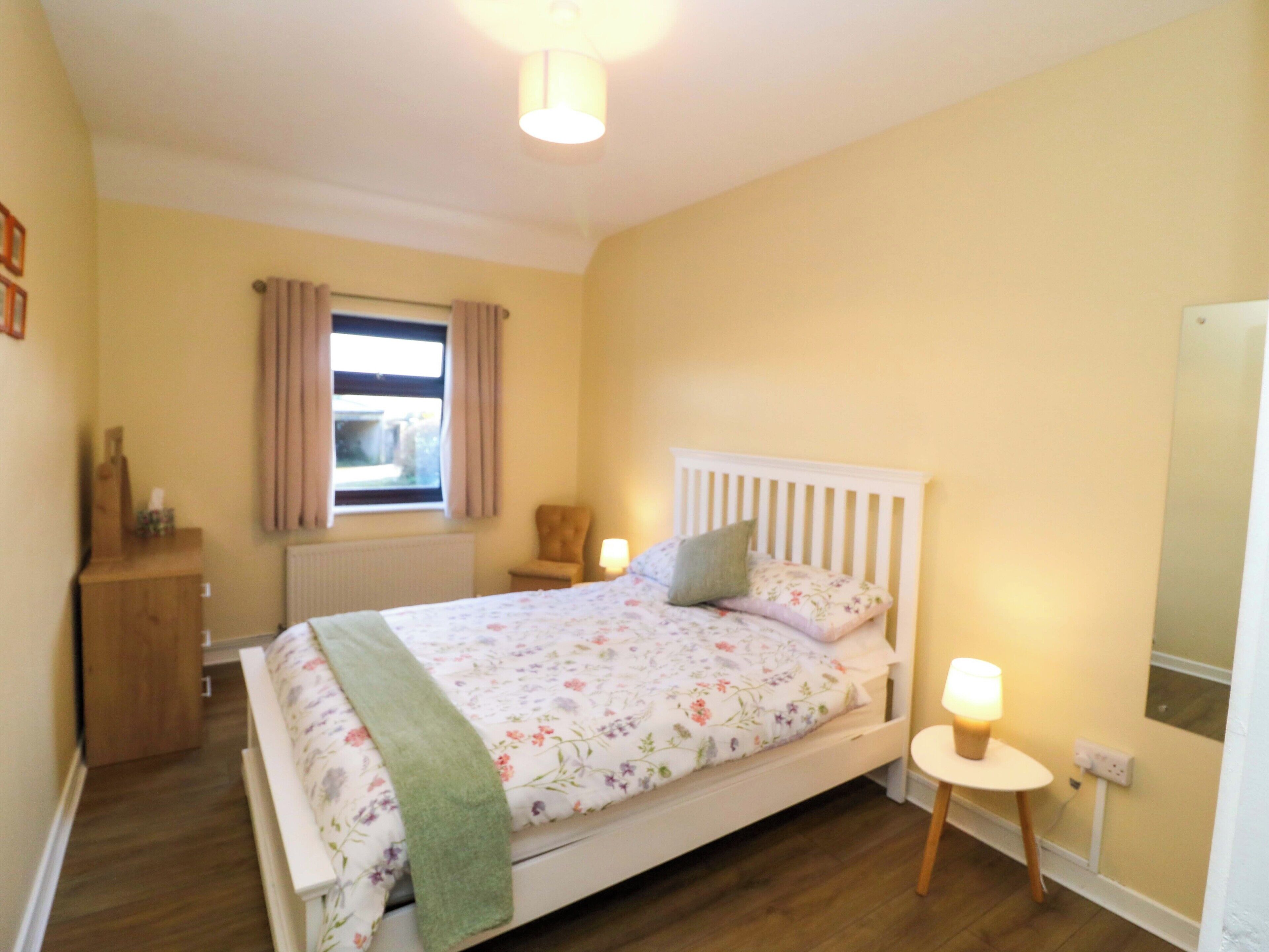 A bedroom with a bed, a window, a chair, a bedside table, and a lamp.