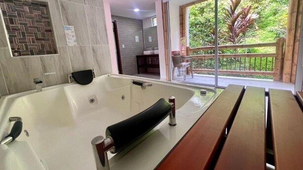 A modern bathroom with a large bathtub, a wooden bench, and a view of greenery through a large window. - Hotel y termales Las Heliconias (Zetaquira)