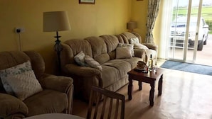 Ein Wohnzimmer mit einer Couch, Sesseln, einem Couchtisch und einer Lampe. Eine Tür führt zu einem Balkon, vor dem ein Auto geparkt ist.
