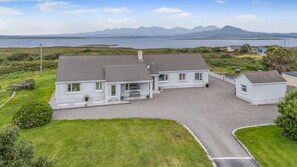 A two-story house with a gravel driveway, a small detached garage, and a view of the sea and mountains in the background. - 3 Bed in Cashel (oc-a32863) (Glynsk Cottages)