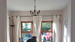 Un comedor con mesa y sillas de madera, dos ventanas con cortinas, una araña y vistas al exterior.