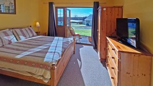 Un dormitorio con cama, televisor, cómoda y vista a una casa y paisaje a través de una puerta.