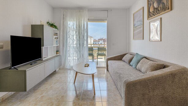 A modern living room with a flat-screen TV, a white coffee table, a beige sofa, and a view of buildings through a window. - Apartment 'Las Conchas' with Sea View, Wi-Fi and Air Conditioning (Cunit)