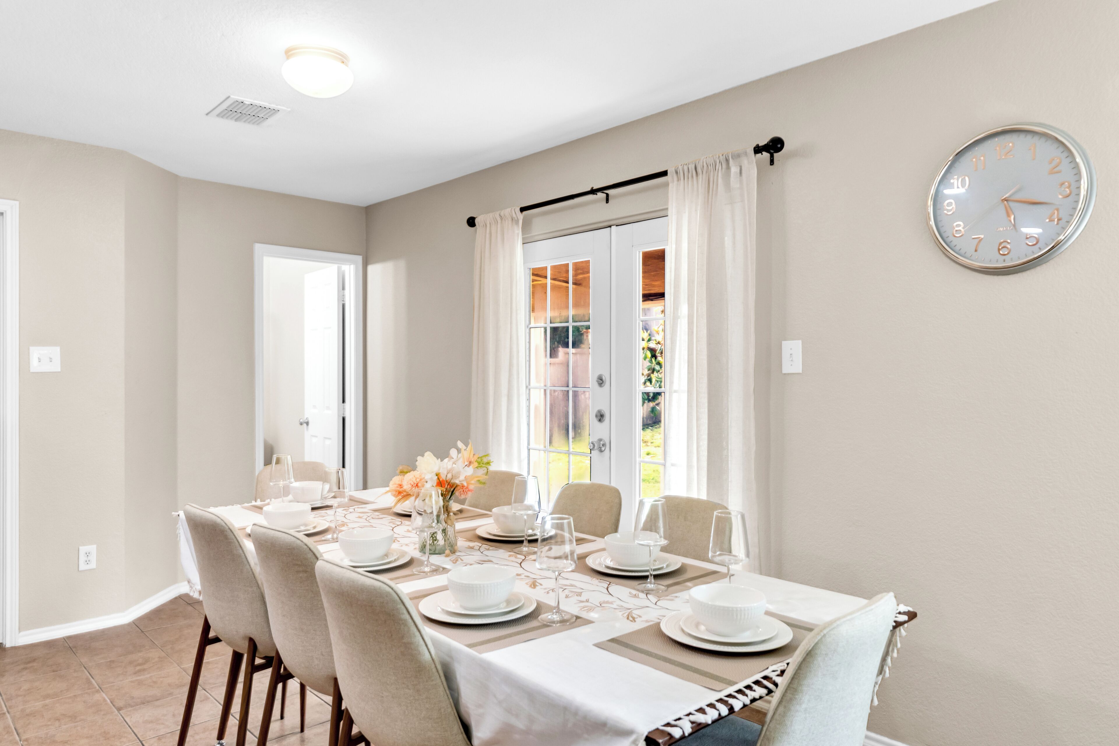 A dining area with a table set for six, a clock on the wall, and a view through a window.