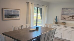 Ein Essbereich mit einem Holztisch, weißen Stühlen, einem gerahmten Bild und Blick durch ein Fenster mit Vorhängen.
