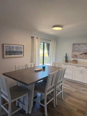 A dining area with a wooden table, white chairs, a framed picture, and a view through a window with curtains. - Cozy 3-bedroom condo in Seascape resort (Miramar Beach)