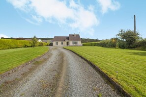 A gravel driveway leading to a two-story house with a sloped roof, surrounded by well-maintained grass lawns. - 3 Bed in Moville (oc-ii07029) (Moville)