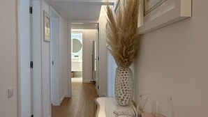 Un couloir avec un mur blanc, un tableau encadré, un vase contenant des plantes séchées et un porte-bougie en verre.