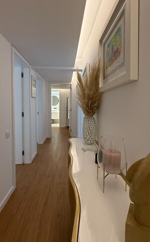 Ein Flur mit weißer Wand, einem gerahmten Gemälde, einer Vase mit Trockenblumen und einem Glas-Kerzenhalter.