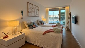 Une chambre avec deux lits, une table de chevet avec une lampe, un téléviseur fixé au mur et une fenêtre donnant sur l’extérieur.