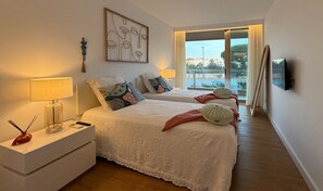 Ein Schlafzimmer mit zwei Betten, einem Nachttisch mit Lampe, einem an der Wand montierten Fernseher und einem Fenster mit Blick nach draußen.