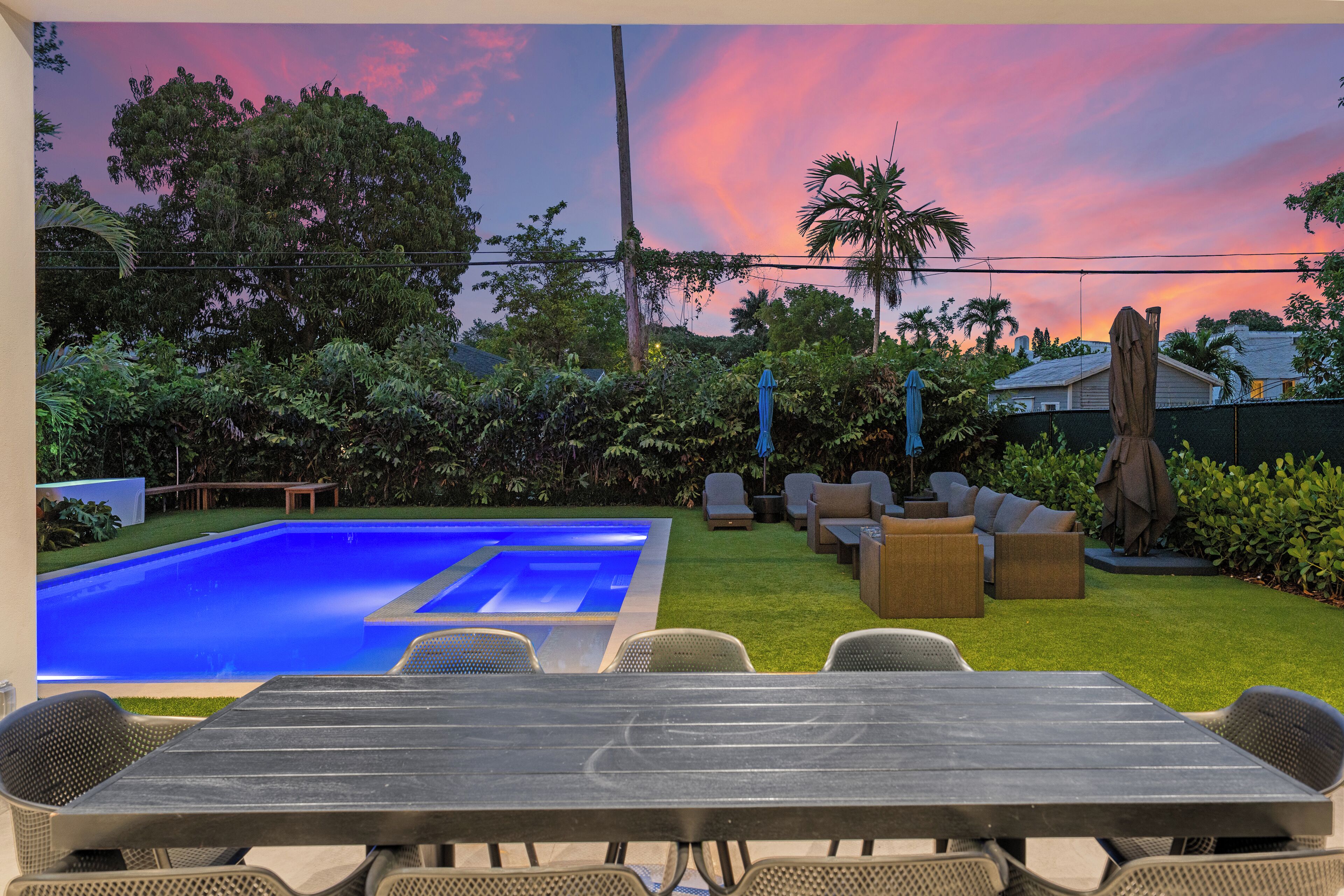 A pool with a blue tint, surrounded by a grassy area with outdoor furniture and a sunset sky.