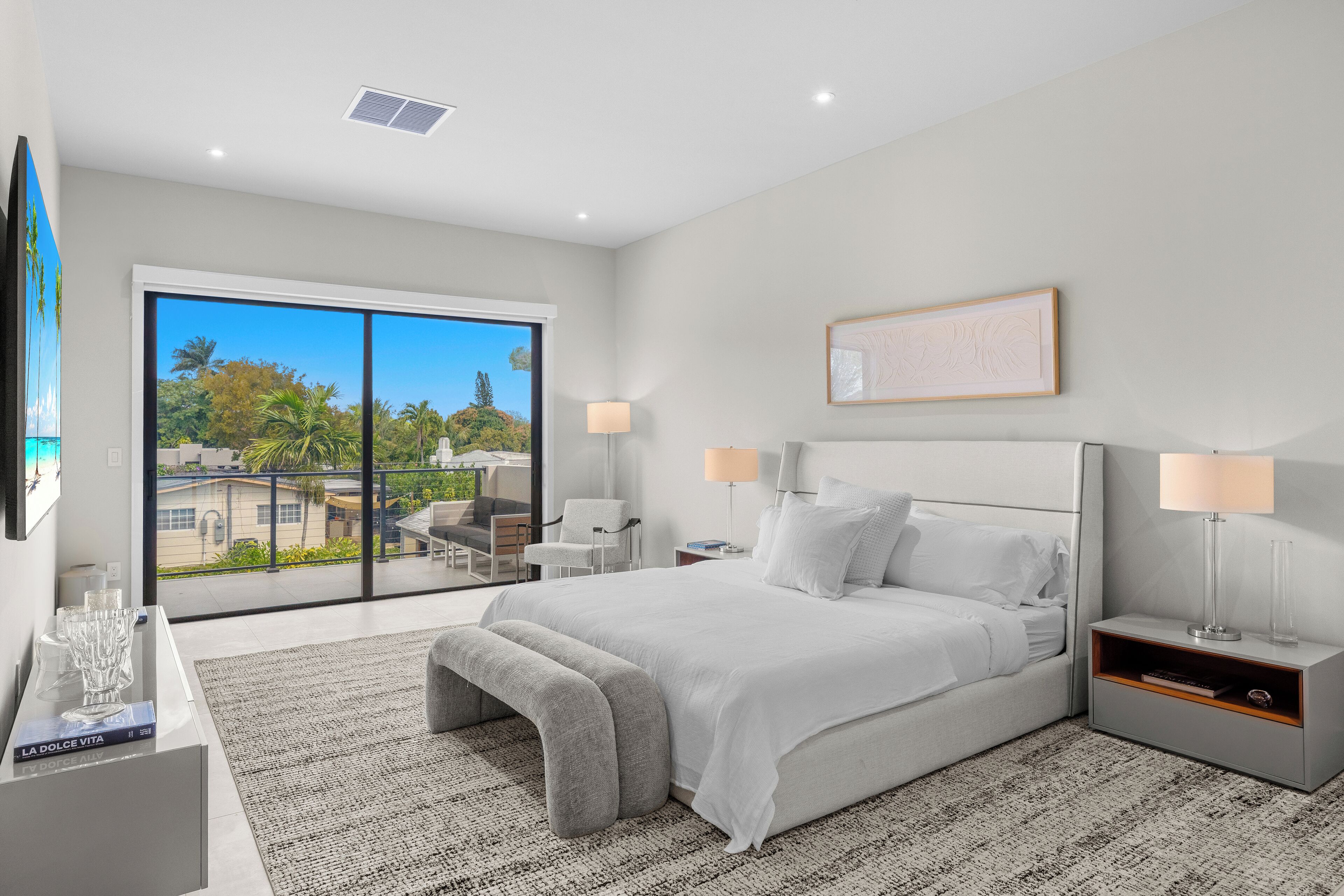 A modern bedroom with a large bed, two bedside tables, a desk with a chair, a large window, and a balcony.