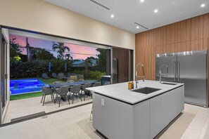 Une cuisine moderne dotée d’un grand îlot central, d’appareils électroménagers en acier inoxydable et offrant une vue sur un jardin avec une piscine et un espace salon extérieur.