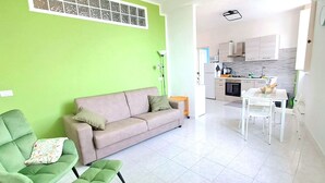 Un salon avec un mur vert, un canapé beige et un coin repas comprenant une table et des chaises blanches.