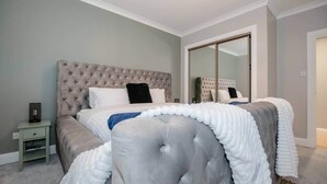 Quarto com cabeceira estofada, cama com roupa de cama branca e cinza, mesa de cabeceira com abajur e um espelho grande.