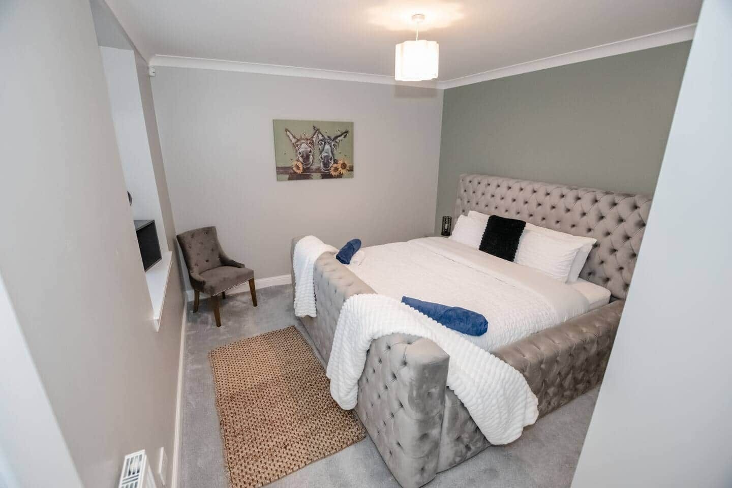 Ein Schlafzimmer mit einem geblümten Kopfteil, einem Bett mit weißer und blauer Bettwäsche, einem Sessel, einem Teppich und einem Wandbild.