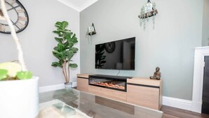 Uma sala de estar moderna com uma TV de tela plana, um rack de madeira, uma mesa de centro de vidro, uma grande planta verde e uma luminária pendente.