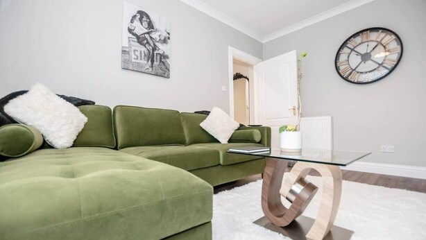 A green sectional sofa with white pillows, a glass coffee table with a wooden base, a wall clock, and a poster on the wall. - The White House East Wing near Edinburgh (Bathgate)