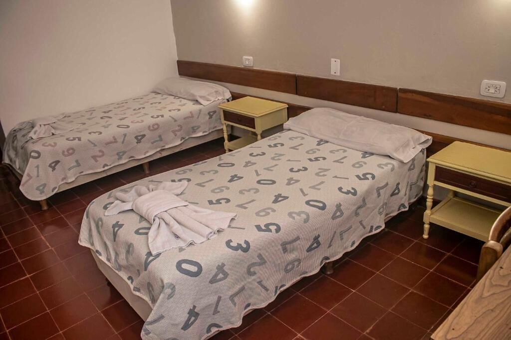 Dos camas individuales con cabeceras y mesitas de noche a juego, piso de baldosas y una pared lisa.
