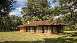 A rustic wooden cabin with a glass-enclosed porch, surrounded by a grassy field and tall trees.