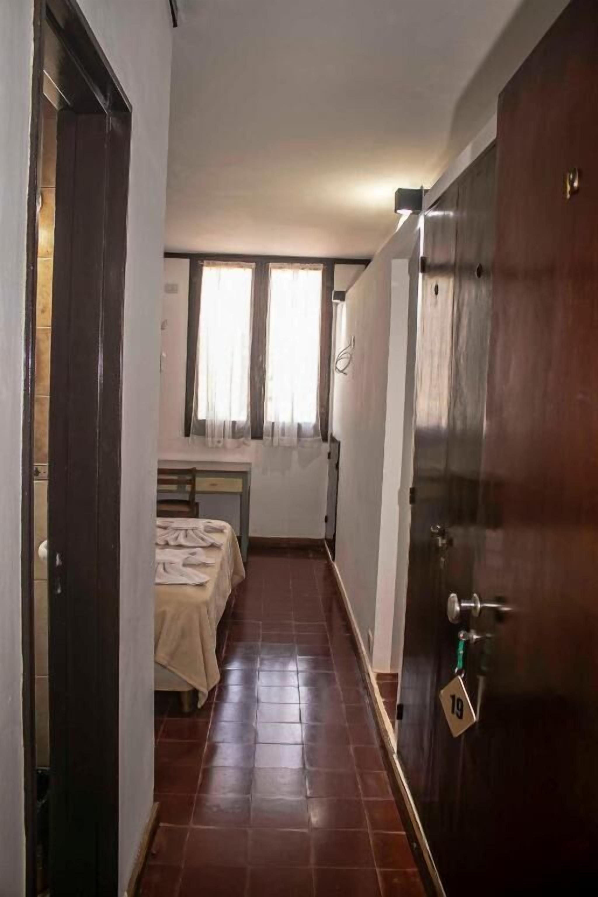 A narrow hallway with a bed and a desk, a door with a number, and a window with curtains.