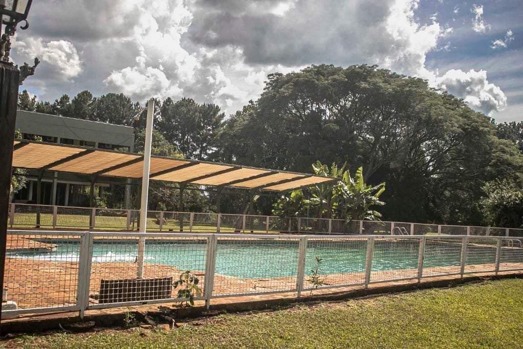 A swimming pool with a fence, a covered area, and trees in the background.