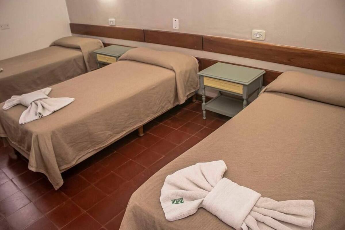 Une chambre d’hôtel avec trois lits simples, chacun équipé d’un oreiller et d’une serviette. Il y a des tables de chevet avec des lampes et une prise de courant au mur.