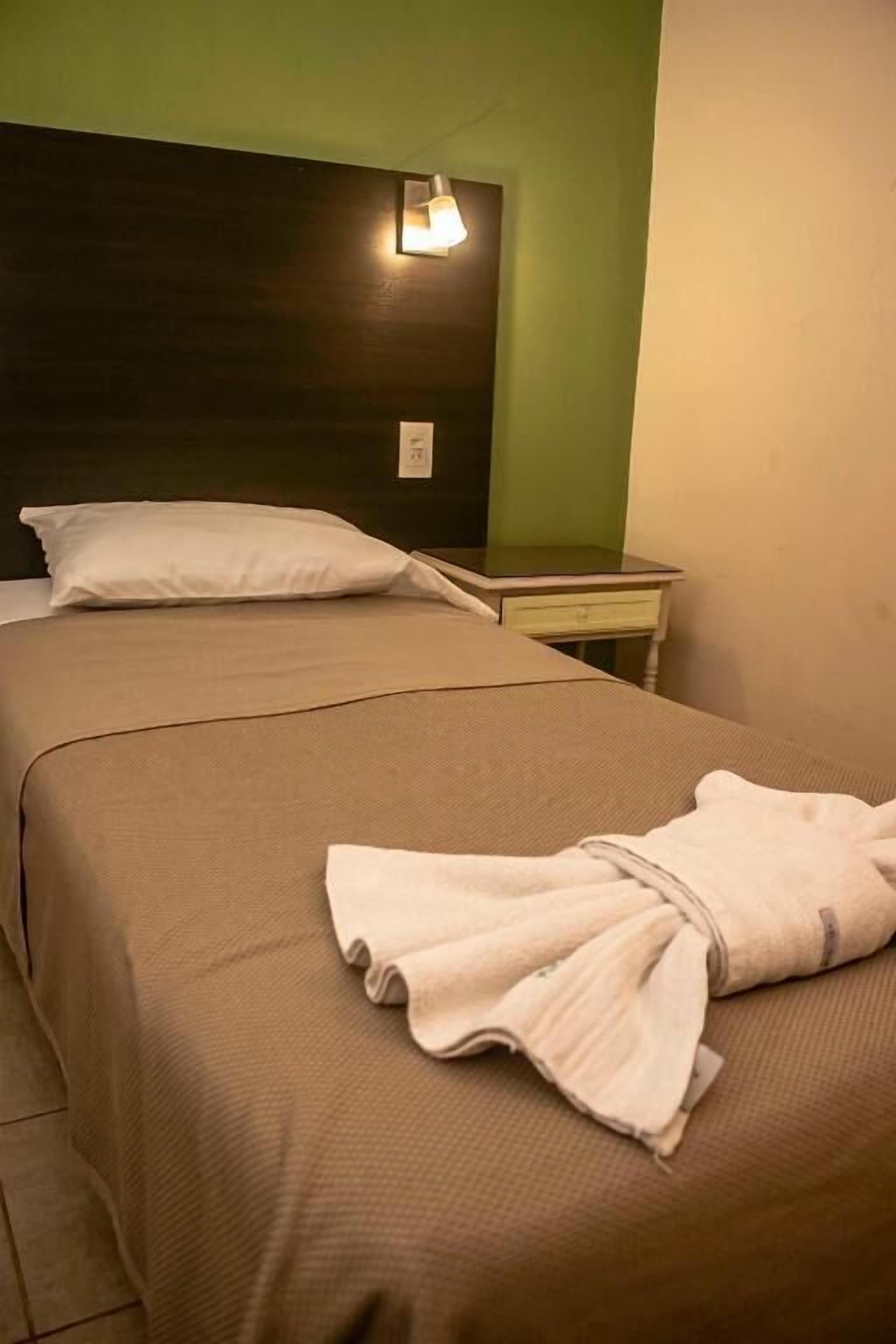 Quarto de hotel com duas camas, uma mesinha de cabeceira, uma luminária e um roupão dobrado na cama.