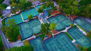 Vue aérienne d’un complexe sportif comprenant plusieurs courts de tennis, entourés d’arbres et de zones de stationnement.