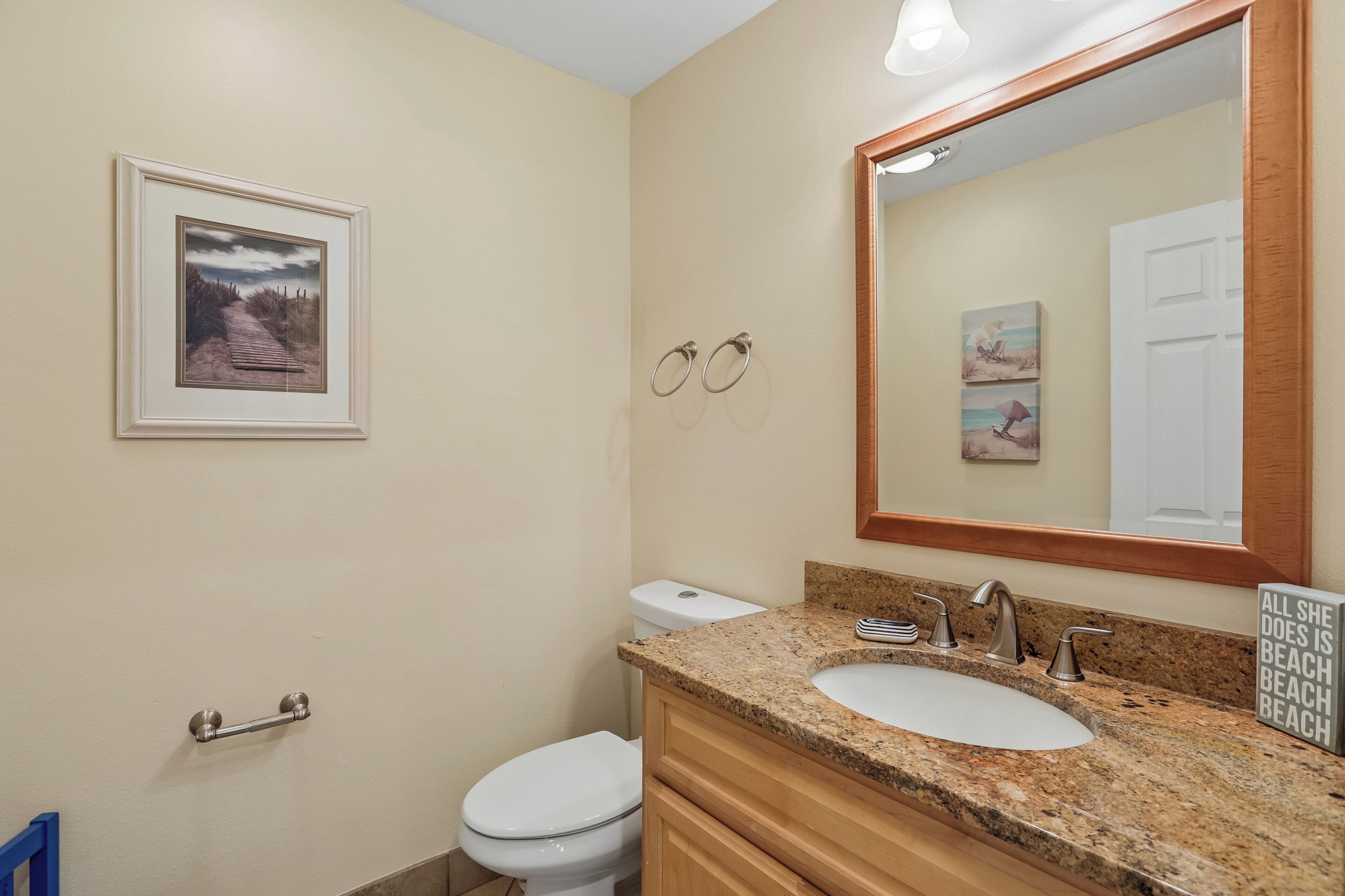 A bathroom with a white toilet, a sink with a granite countertop, a wooden vanity, and a large mirror. There is a framed picture on the wall and a towel rack with two towels.