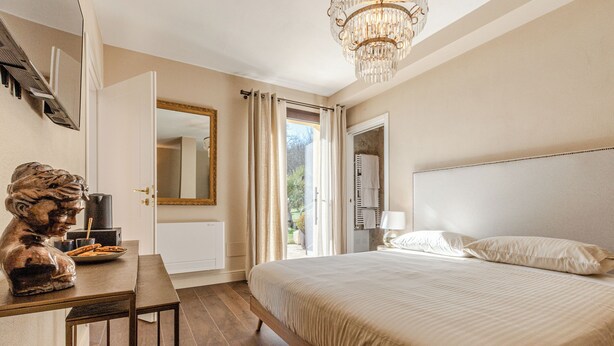 Une chambre à coucher avec un lit, un bureau orné d’une statue, un lustre et une vue sur l’extérieur, visible à travers la porte ouverte.