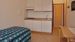 A compact kitchen with a sink, cabinets, and a stove. There is a bed with a patterned bedspread and a wooden table with a chair. - Functional and Comfortable Seafront Studio (San Michele al Tagliamento)