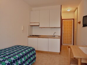 Eine kleine Küche mit Spüle, Schränken und einem Herd. Ein Bett mit einem gemusterten Überwurf und ein Holztisch mit einem Stuhl.