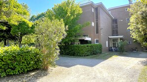 A building with a brick facade, a covered entrance, and a gravel courtyard surrounded by greenery. - Functional and Comfortable Seafront Studio (San Michele al Tagliamento)