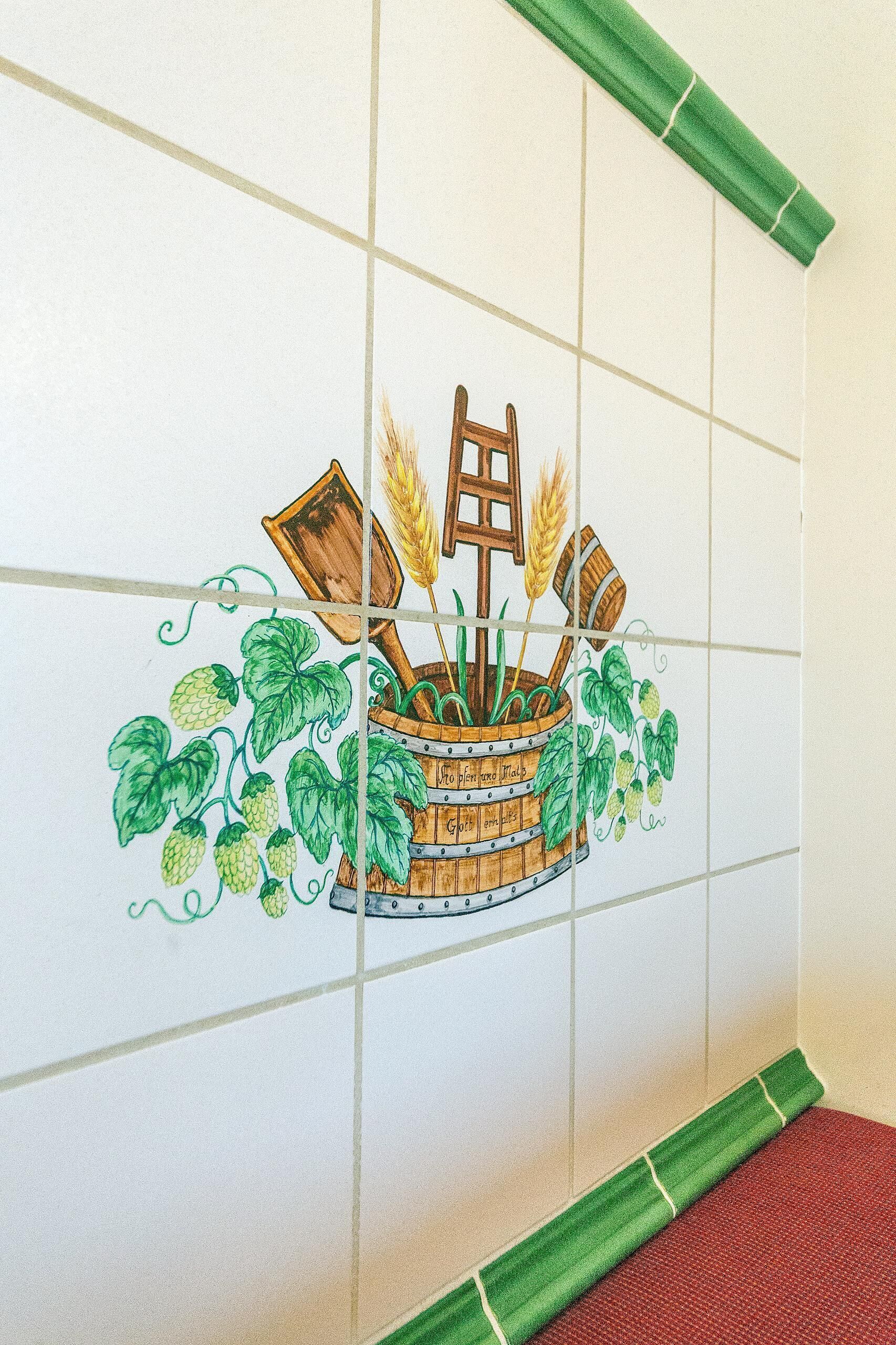 Een betegelde muur met een decoratieve afbeelding van een houten vat, graanhalmen en hopbellen.