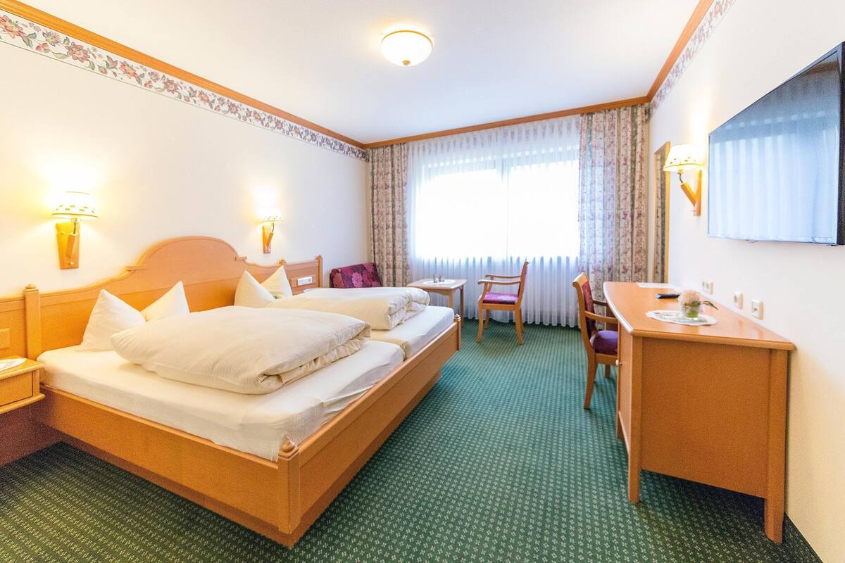 A hotel room with a double bed, a desk, two chairs, a TV, and a window with curtains.