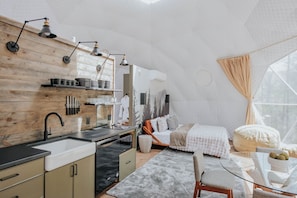 A modern interior with a kitchen, dining area, and bedroom, featuring wooden walls, a large window, and a unique dome ceiling. - Glamp Blue Ridge - Dome 2 Brunson (Gilmer County)