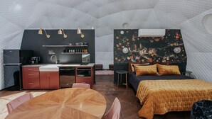 Una habitación compacta y moderna con kitchenette, una cama con cabecera floral, una mesa de comedor y un techo geométrico único.