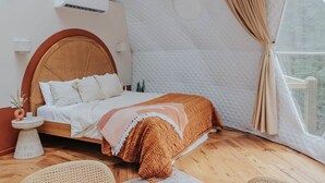 A bedroom with a bed, a round table, wicker chairs, a bedside table with a vase, and a window with curtains.