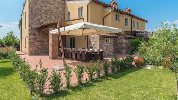 Ein zweistöckiges Steinhaus mit einer Terrasse, auf der ein Esstisch und Stühle stehen, umgeben von einem gepflegten Garten.