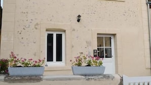 Un edificio de dos pisos con fachada color beige, dos ventanas en la planta baja y un pequeño balcón con plantas en macetas.