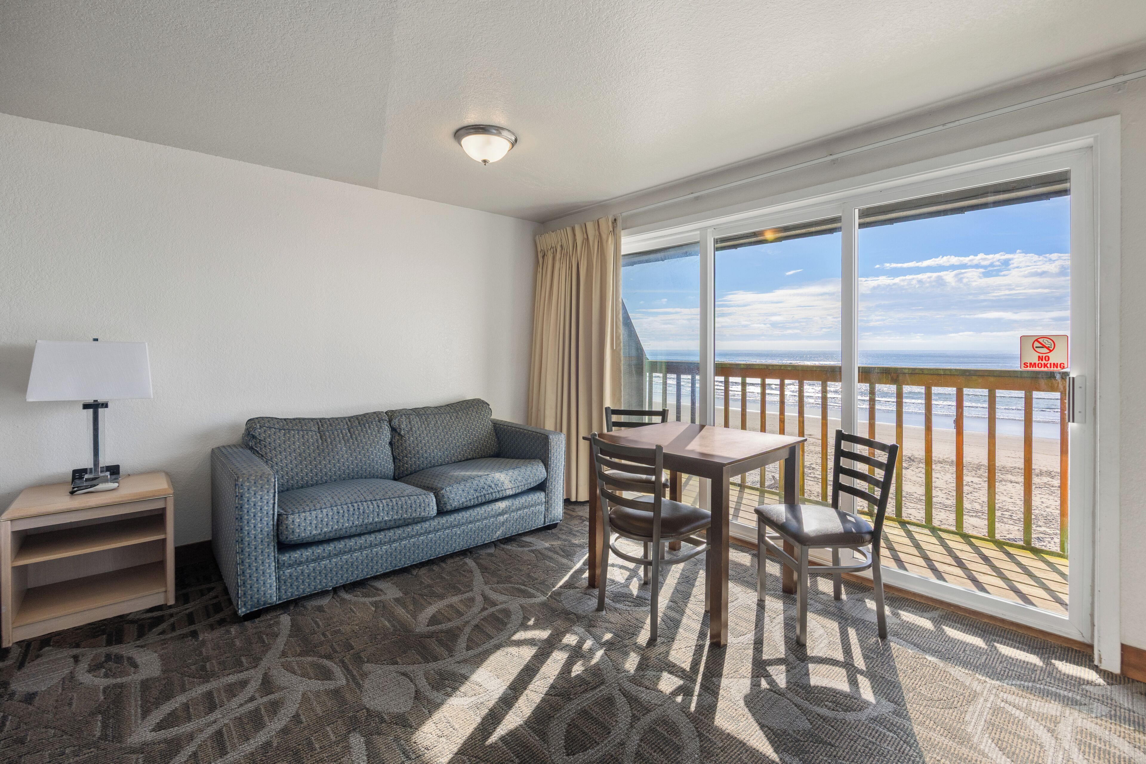 A hotel room with a sofa, a table with chairs, a lamp, and a view of the beach through a large window.