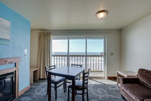 A room with a dining table, chairs, a sofa, a fireplace, and a large window with a view of the beach. - Surfside Resort (Rockaway Beach)