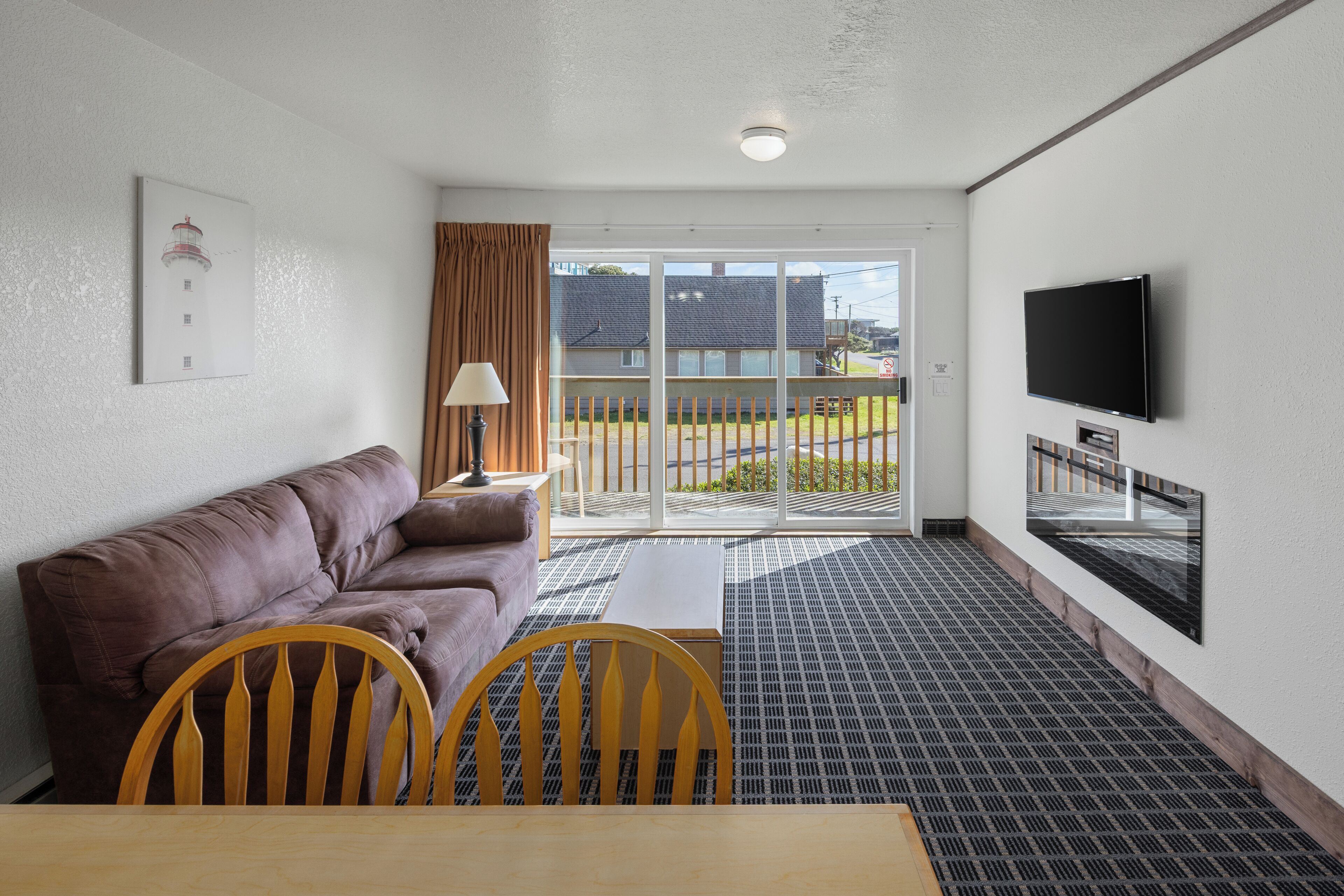 A hotel room with a brown sofa, a small table, two wooden chairs, a TV mounted on the wall, and a sliding glass door leading to an outdoor area.