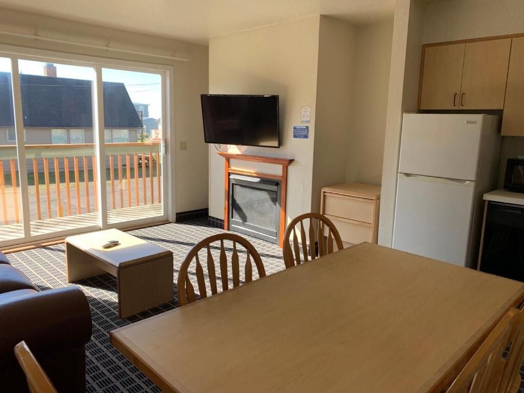 Una habitación con una mesa de comedor, sillas, un televisor sobre la chimenea, un refrigerador y un ventanal corredero que da a un balcón.