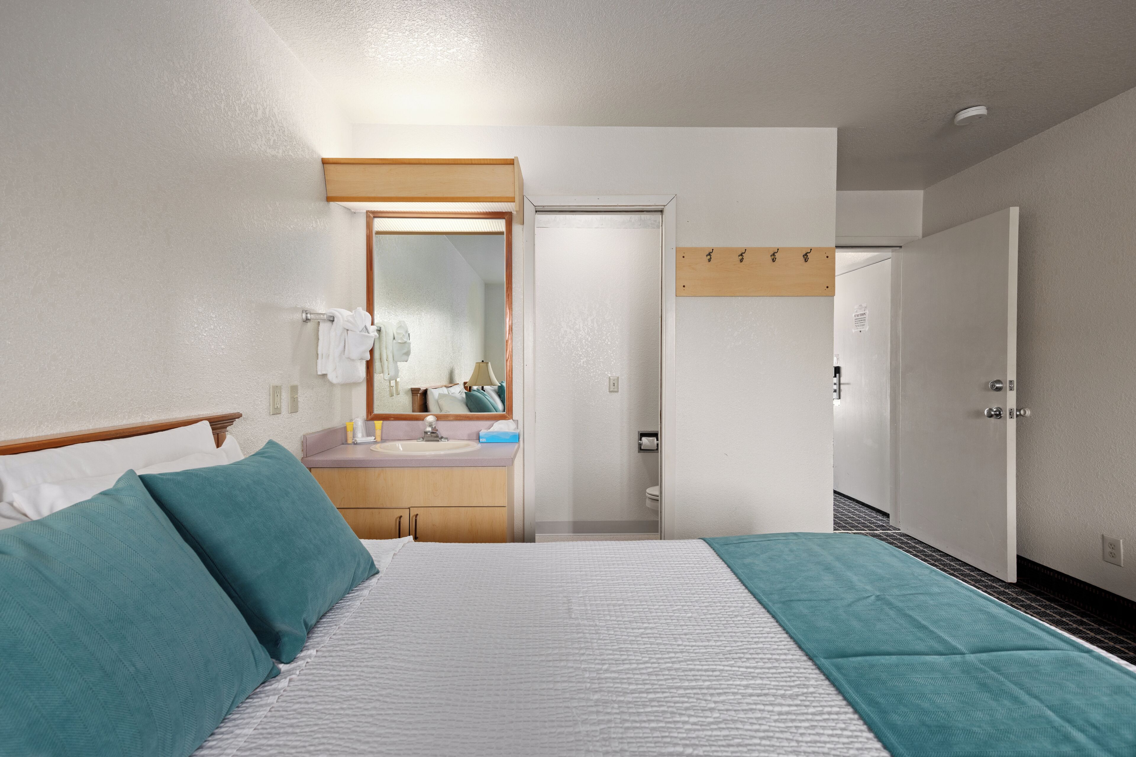Una camera d'albergo con un letto, un comodino, uno specchio, un bagno con lavandino e toilette, e una porta che dà su un'altra stanza.