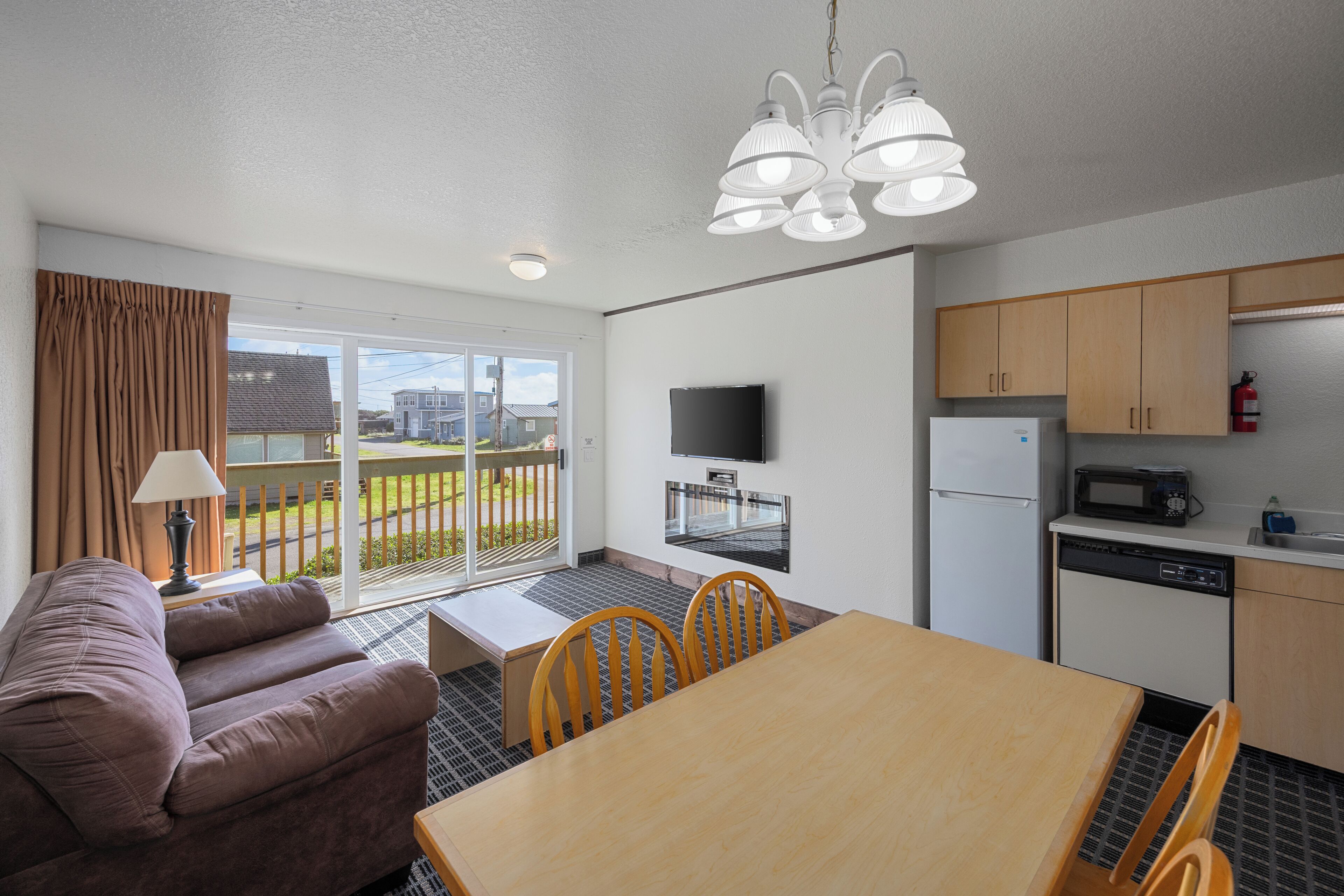 A modern hotel room with a sofa, a dining table, a kitchen area with appliances, and a large window with a view.